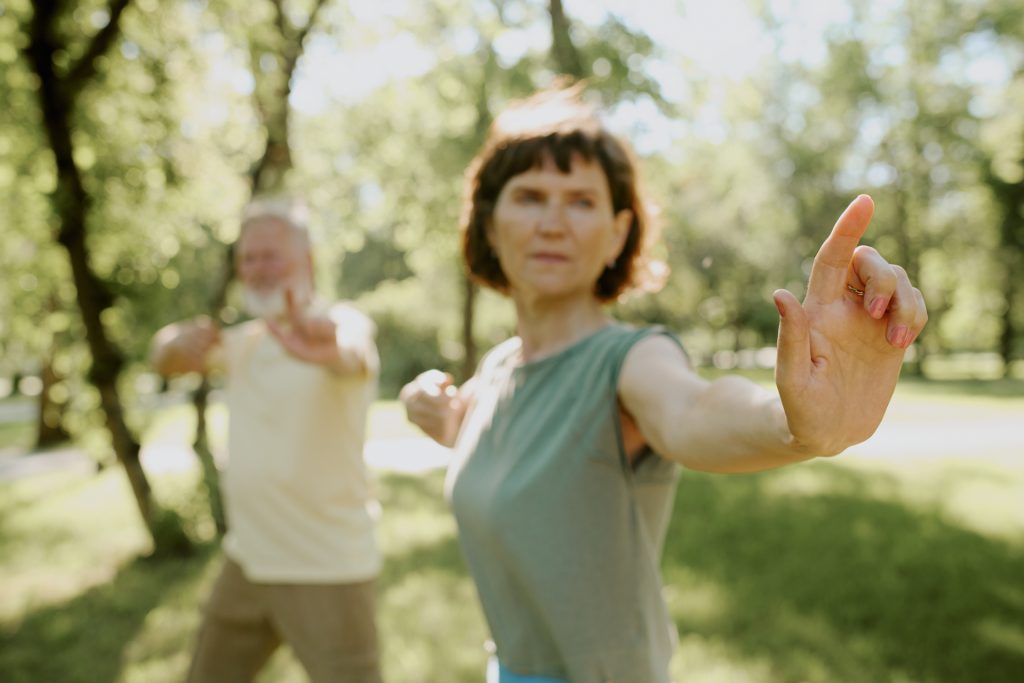 Ältere brünette Frau, die in einer Qigong-Pose steht, mit ihrem grauhaarigen Mensch unscharf im Hintergrund.