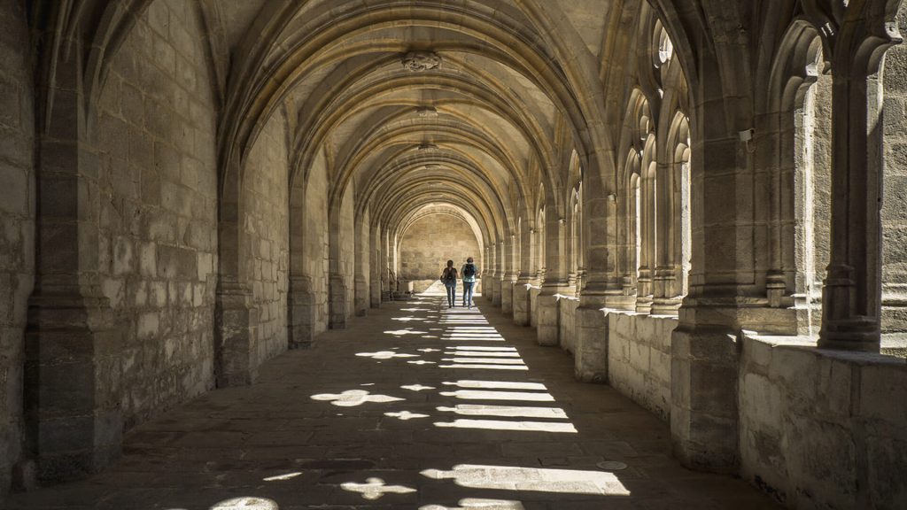 Zwei Personen gehen durch einen historischen, gewölbten Kreuzgang mit Licht- und Schattenspiel.