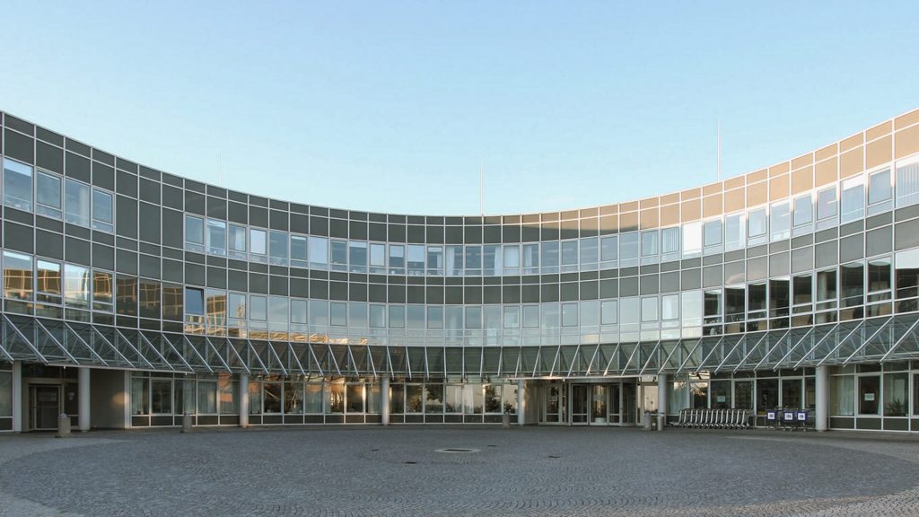 Gebogene Glasfassade eines modernen Gebäudes vor blauem Himmel, mit gepflastertem Vorplatz.