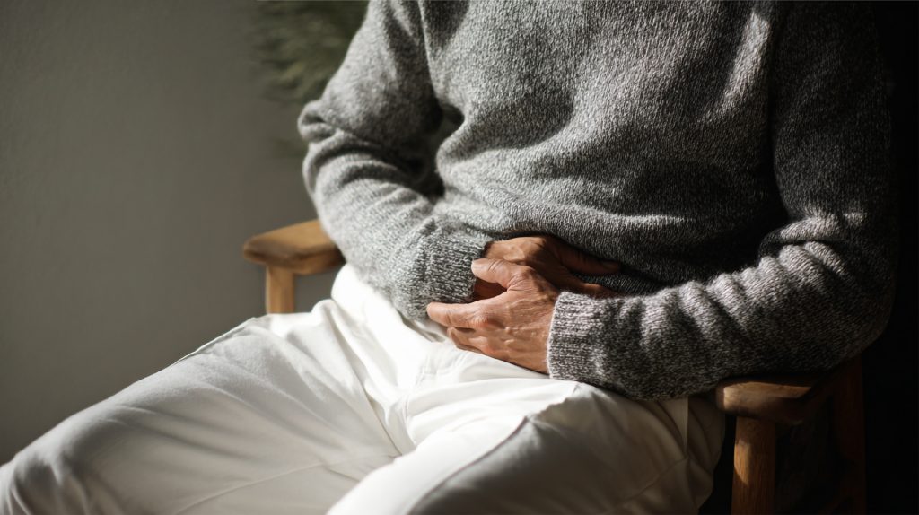 Person sitzt mit Händen auf dem Bauch, trägt grauen Pullover und weiße Hose, Tageslicht erhellt den Raum.