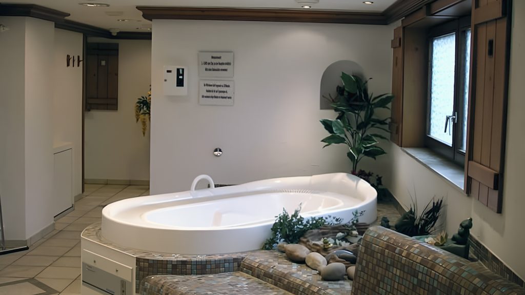 Wellnessbereich mit großer, weißer Badewanne, umgeben von Pflanzen und dekorativen Steinen, neben einem Fenster.